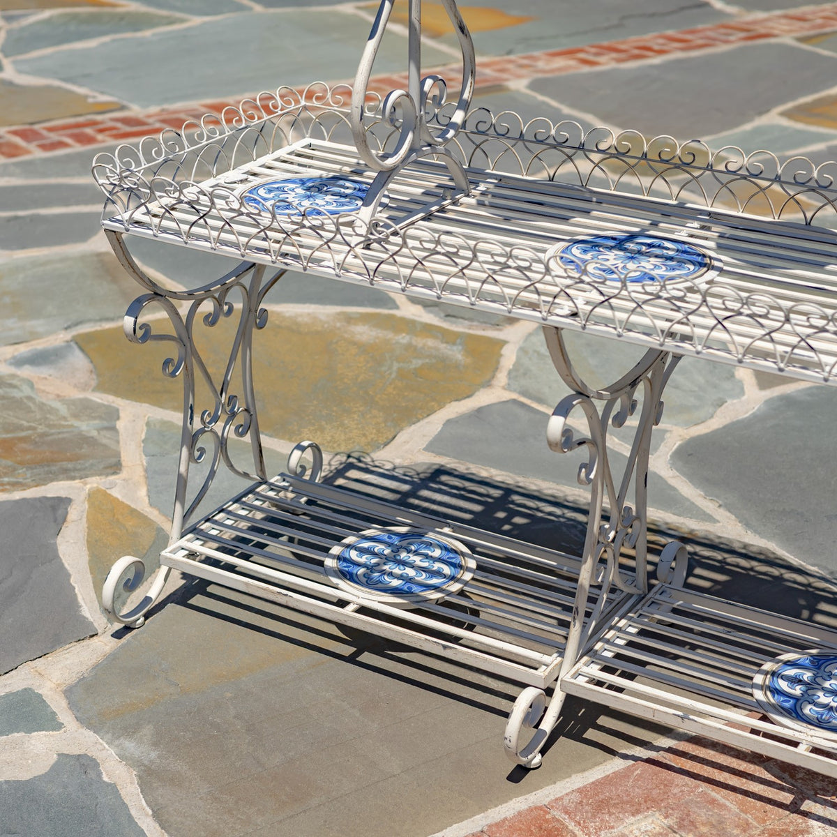 White scrollwork display stand with blue mosaic insets on its slatted tiers, viewed against a stone patio.