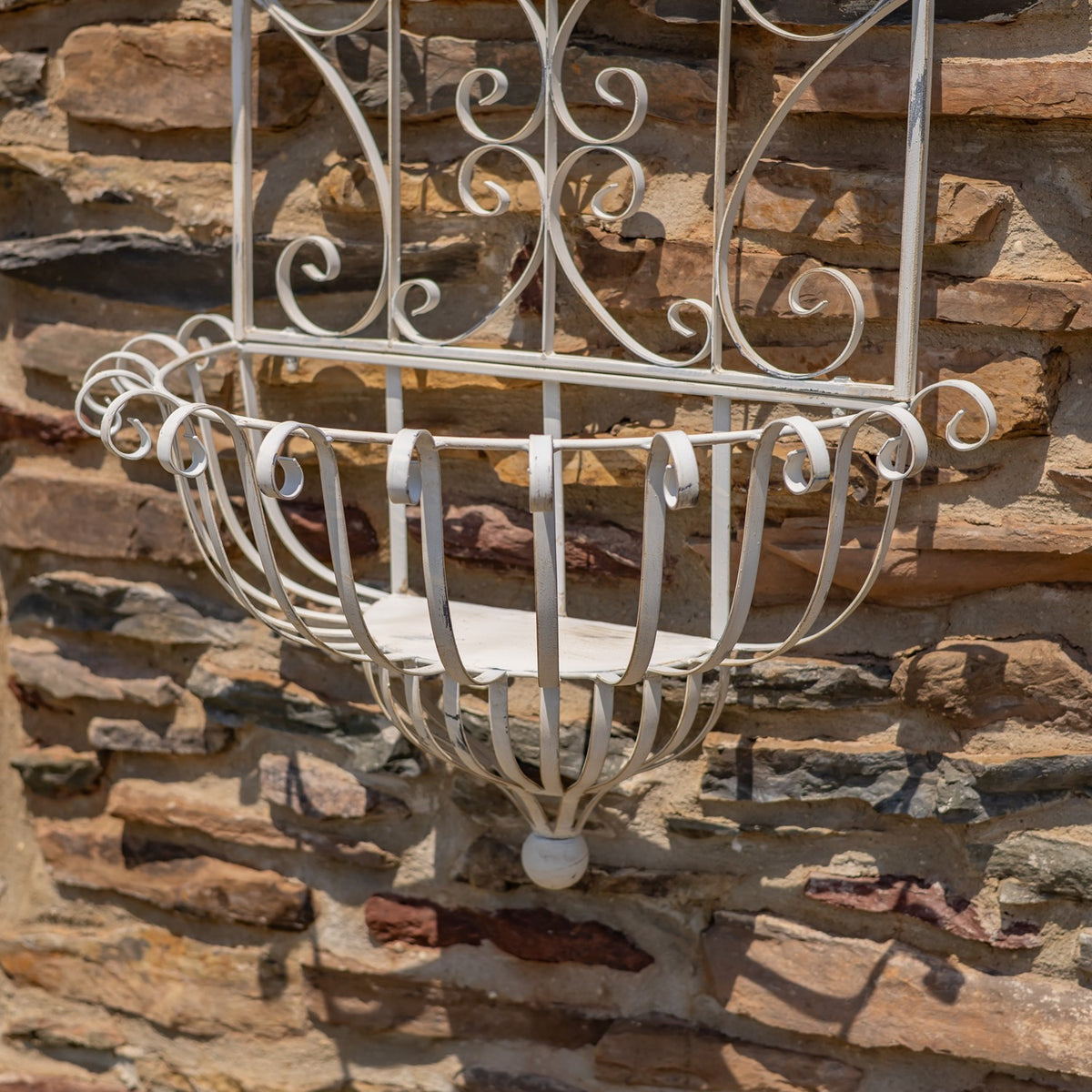 Curved white metal planter basket hangs from an ornate scrollwork frame against a stone wall.