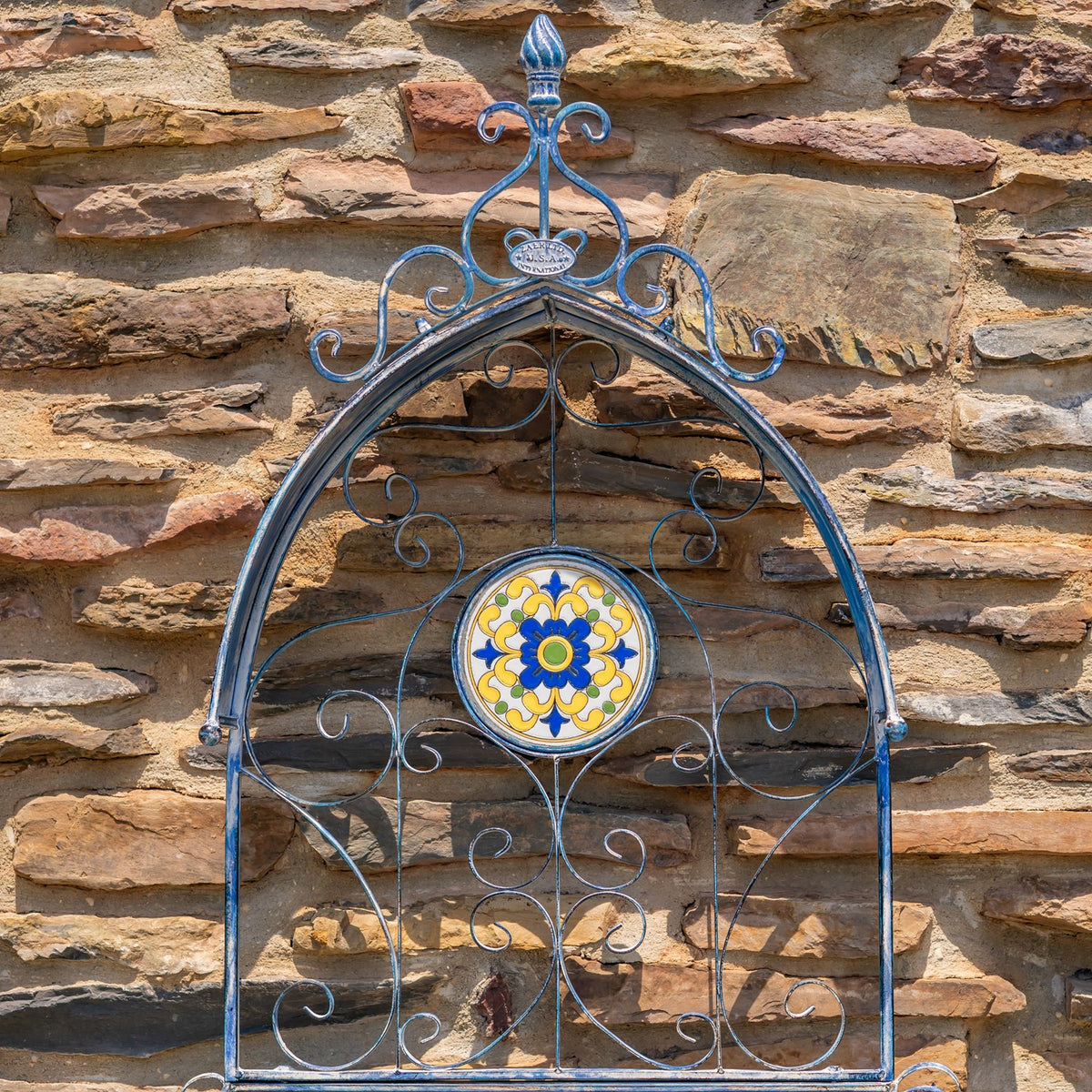 Blue wrought-iron wall planter with curled scrollwork and a round floral mosaic against a stone wall.