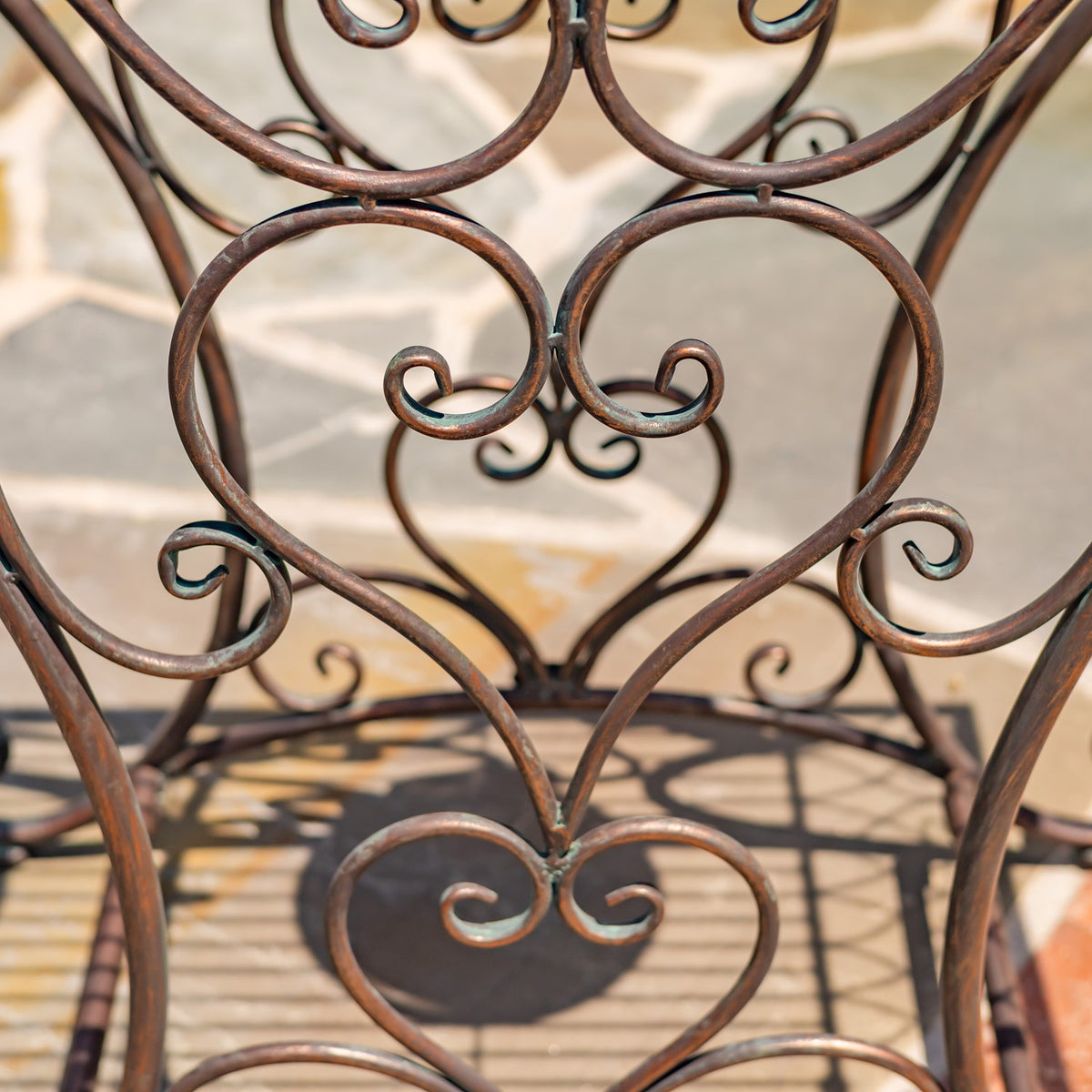 Close-up of a table’s curved bronze-toned iron scrollwork against a sunlit tiled patio.
