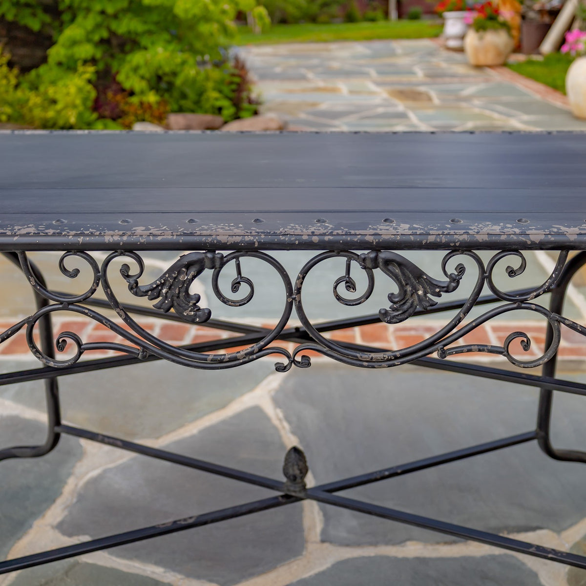 Dark metal dining table with ornate scrollwork beneath the tabletop, shown outdoors.