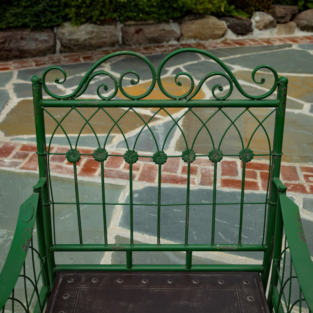 Green metal chair with ornate scrollwork back and a dark brown seat on a stone patio