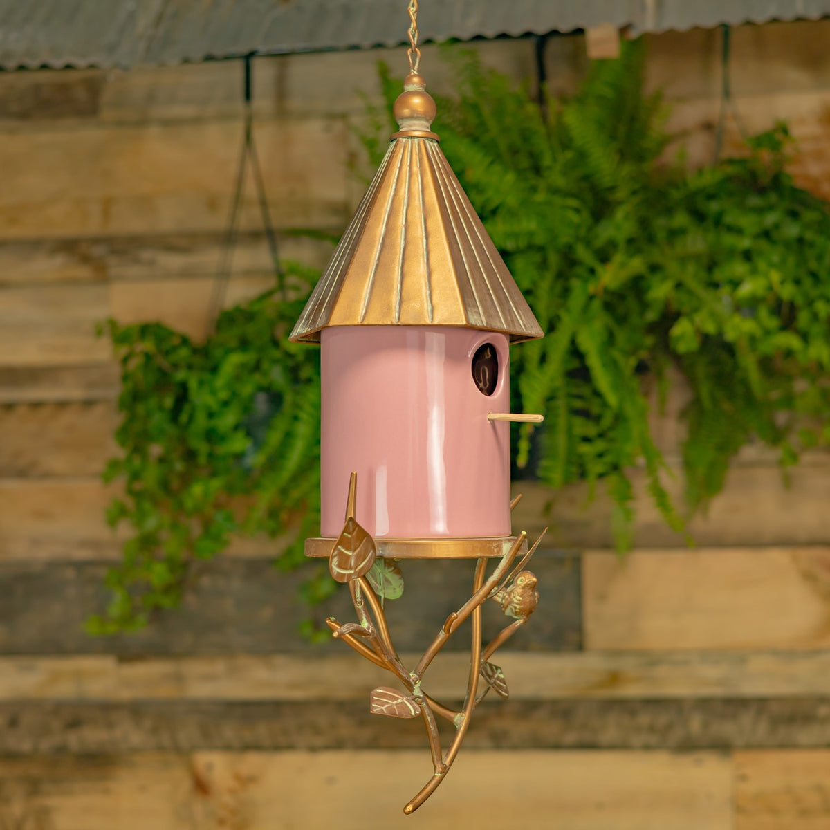 Pink birdhouse with a gold-toned roof and branch-like metal base hangs against a leafy garden backdrop.