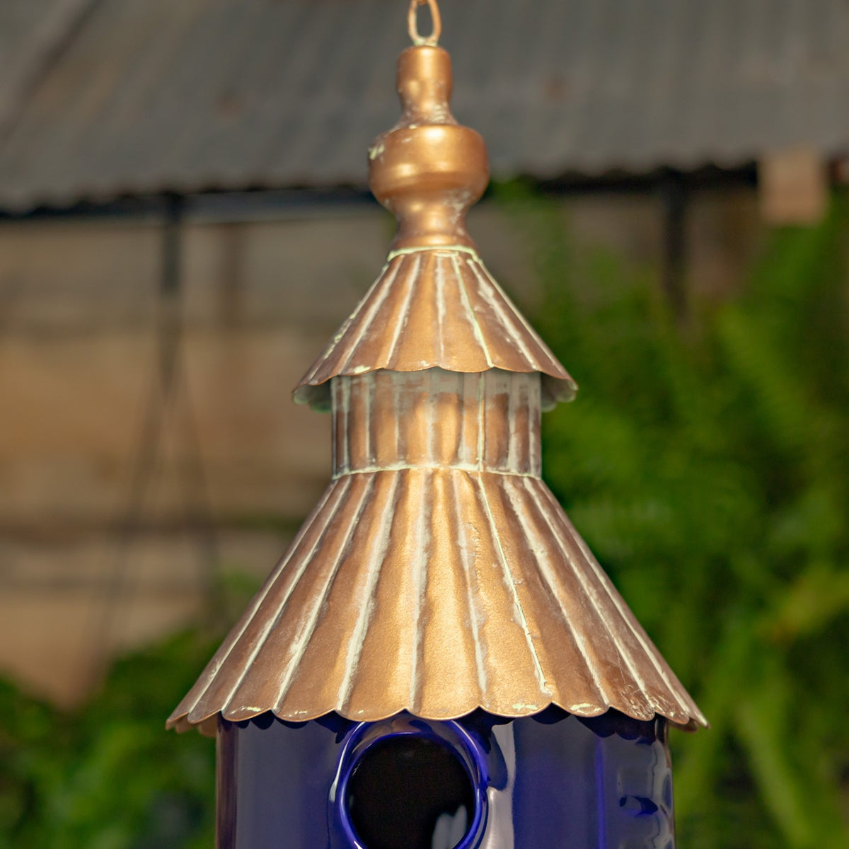 Close view of the birdhouse’s bronze-toned tiered roof and round entrance against a garden background.