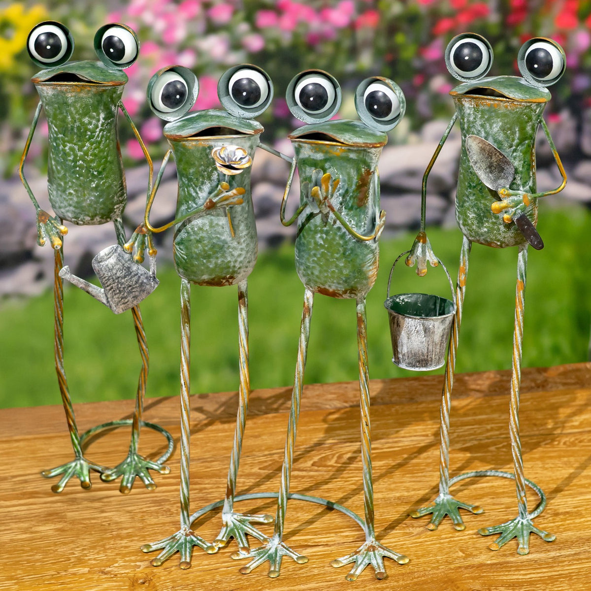 Tall green metal frog figures stand on a wooden surface, some holding small metal pots, with flowers behind them.