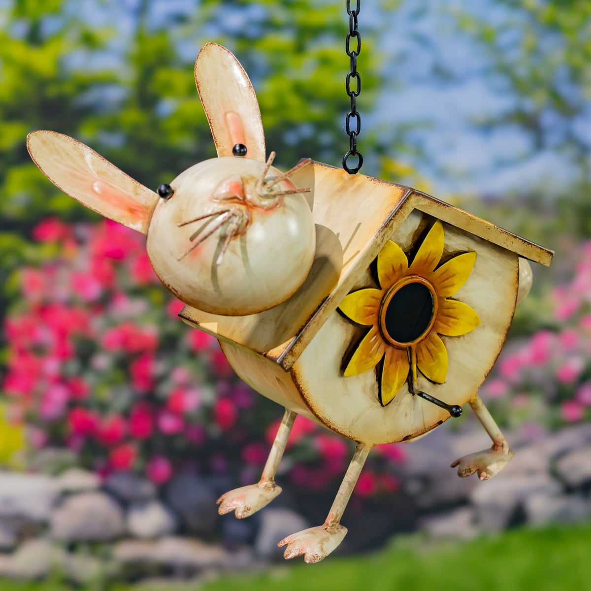 A hanging metal birdhouse shaped like a rabbit, with a sunflower on its body, against a garden backdrop.