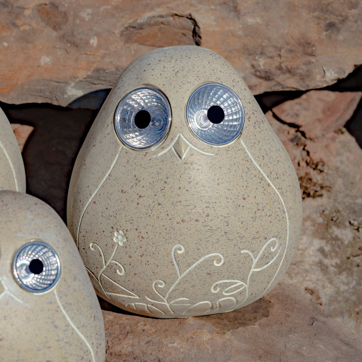 A speckled beige bird ornament with reflective round eyes and white floral linework sits among rocks.