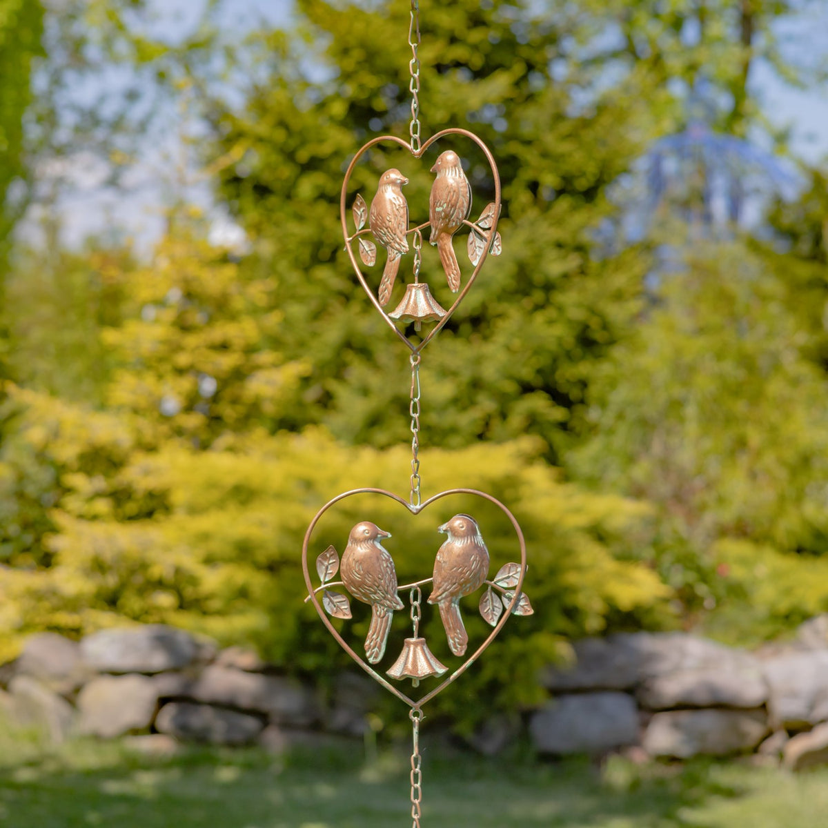 A copper-colored rain chain with two heart-shaped bird ornaments hangs in front of a garden wall.
