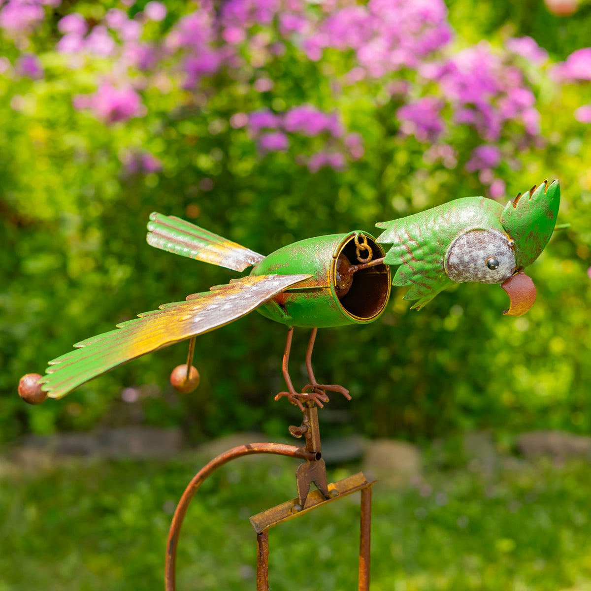 Green metal bird stake shown in side view against pink garden flowers