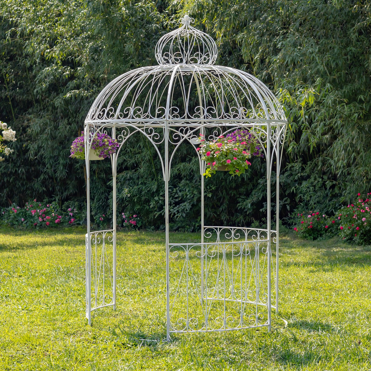 White round iron gazebo with ornate scrollwork and hanging flower baskets on a garden lawn.
