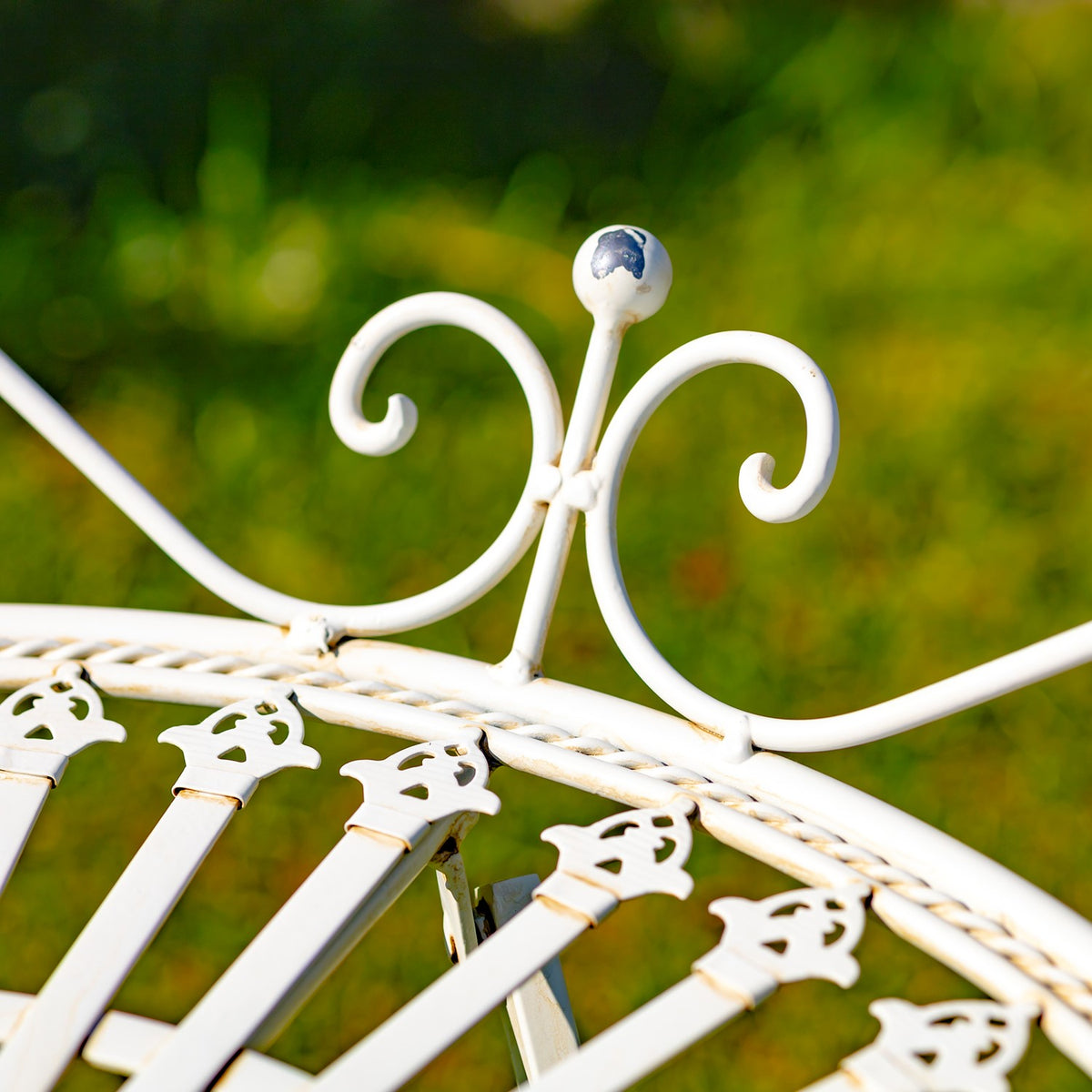 Curled white metal ornament rising from the table rim against a blurred green garden background.