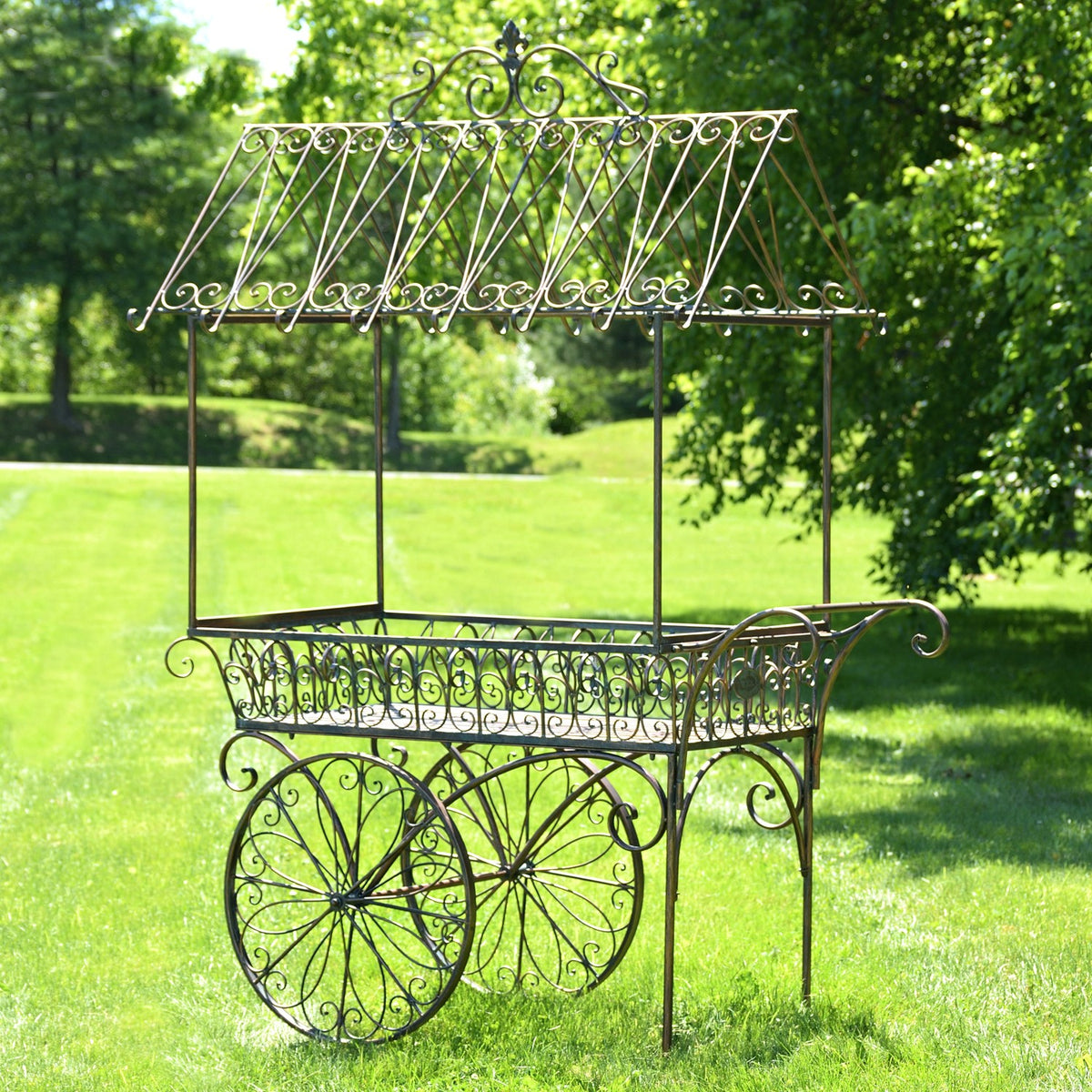 Empty black iron flower cart with scrollwork sides and large spoked wheels on a lawn.