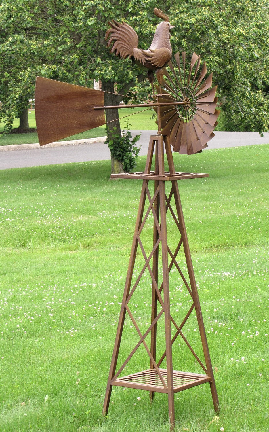 Tall rust-brown metal windmill with a rooster ornament and broad tail, standing on a lawn beneath trees.