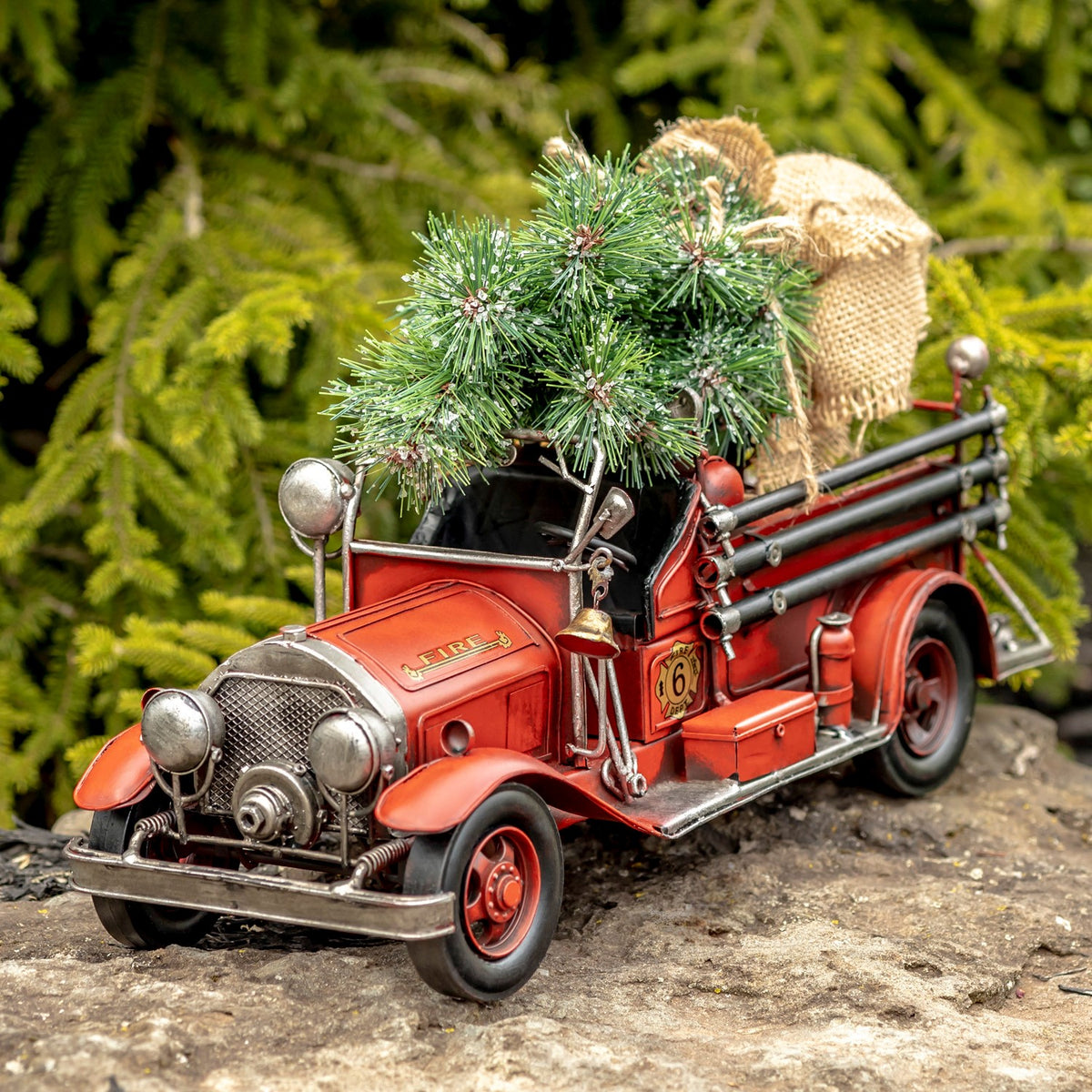 Red vintage-style fire truck with an open cab, evergreen tree, and burlap-wrapped bundle on its rear.