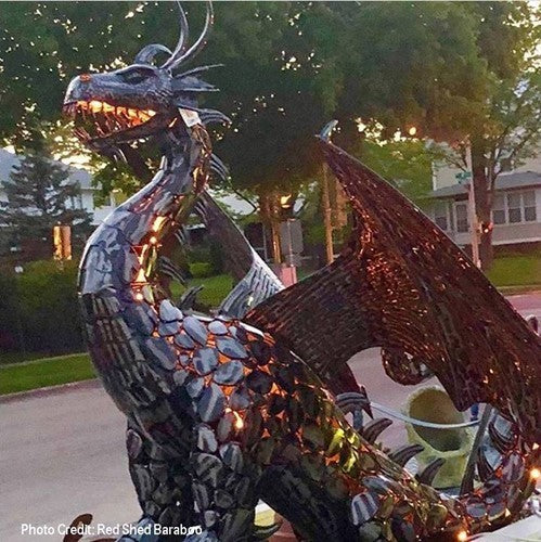 Metal dragon statue glowing with warm lights along its mouth and body, with trees and houses in the background.