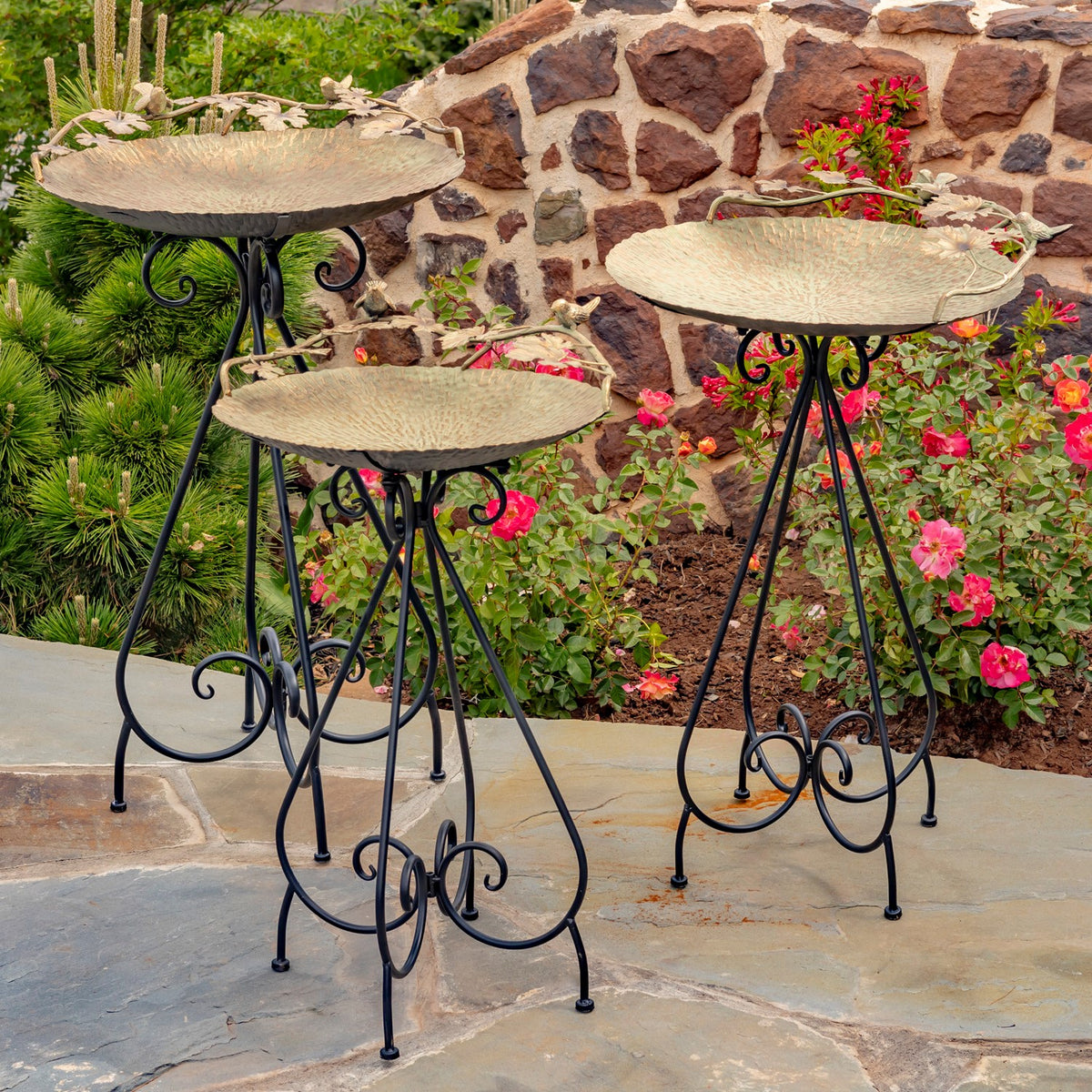 Three metal birdbaths on decorative stands arranged among flowering plants on a stone patio.
