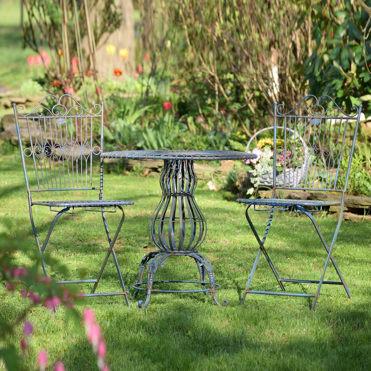 Two ornate metal chairs flank a round pedestal table on a grassy garden lawn.