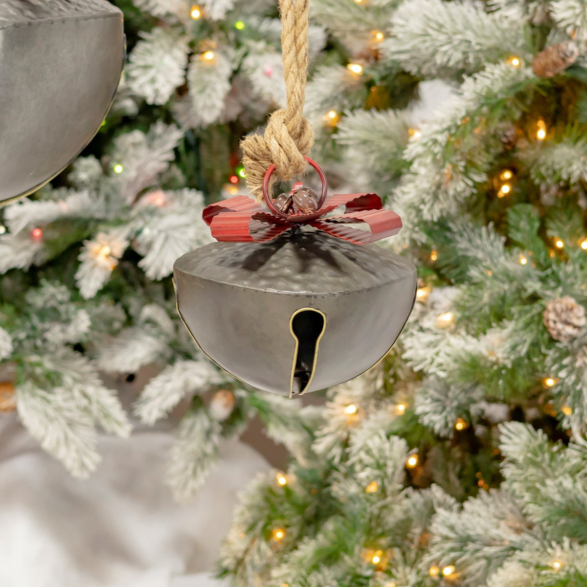 Smaller rounded metal bell with a red bow hanging among snow-dusted evergreen branches.