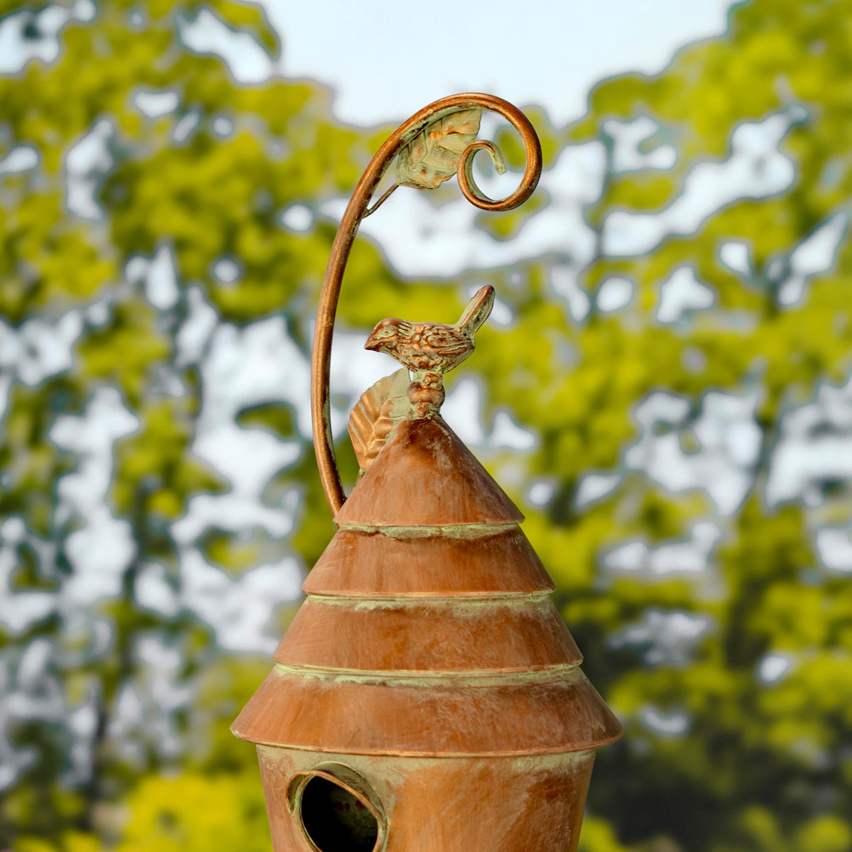 Copper-toned birdhouse with a round entrance, tiered roof, bird ornament, and curled leafy stake top.