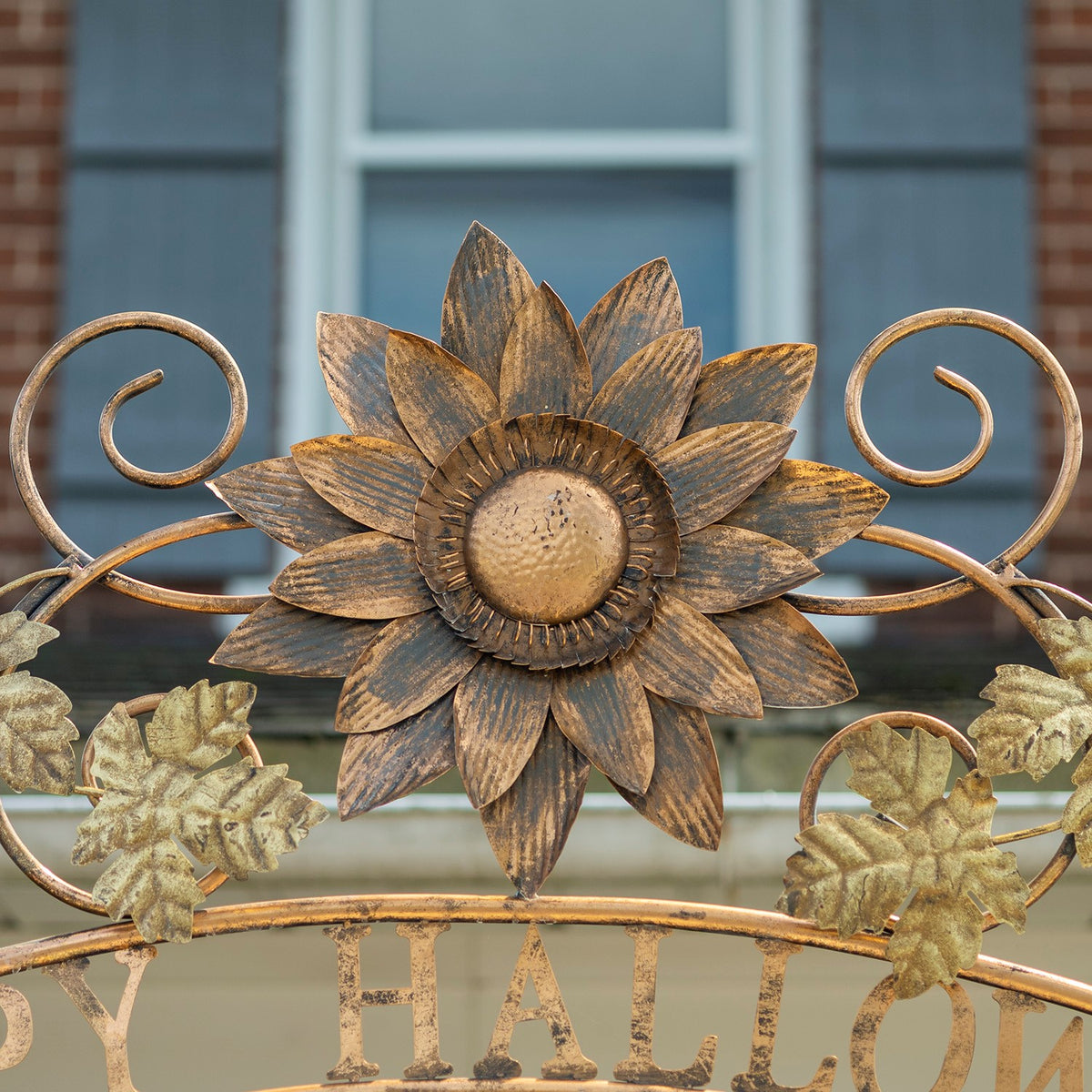 Metal sunflower and curled scrollwork decorate the arch, with pale leaves and partial lettering below.