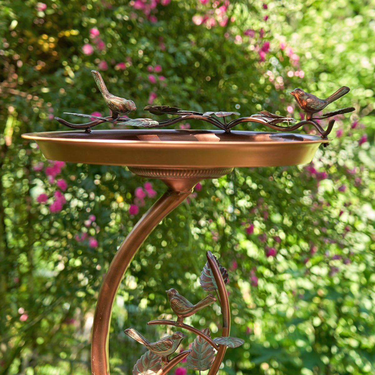 Shallow copper-toned birdbath on a curved branch-style stand, with small bird figures around the rim and base.