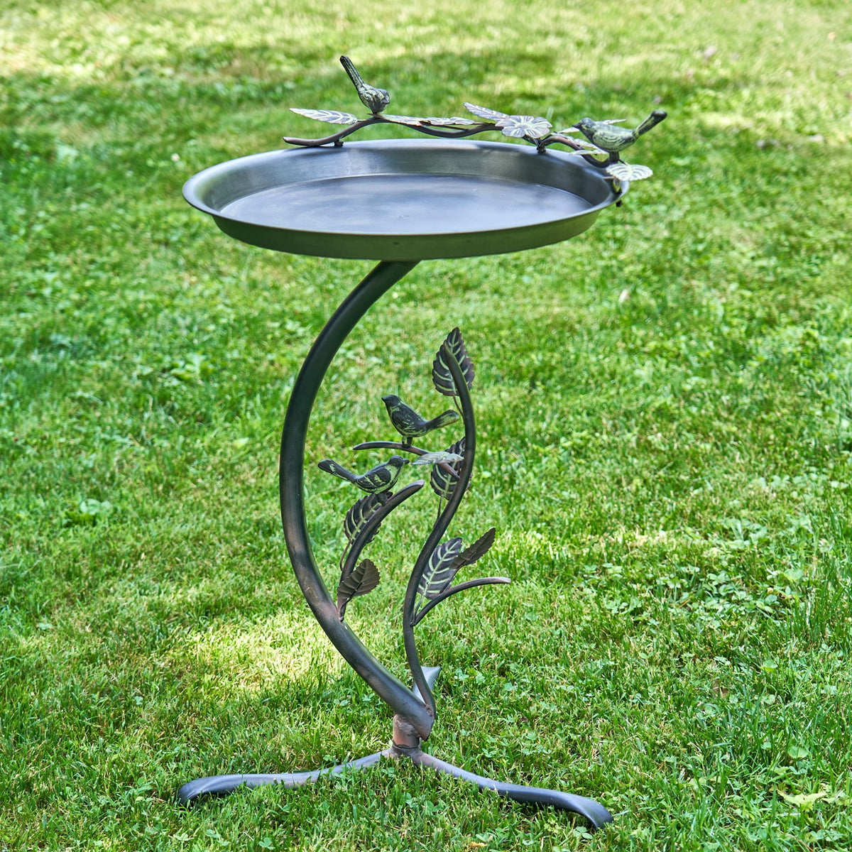 Metal birdbath with a decorative branch stand and curled feet, shown on a grassy lawn.