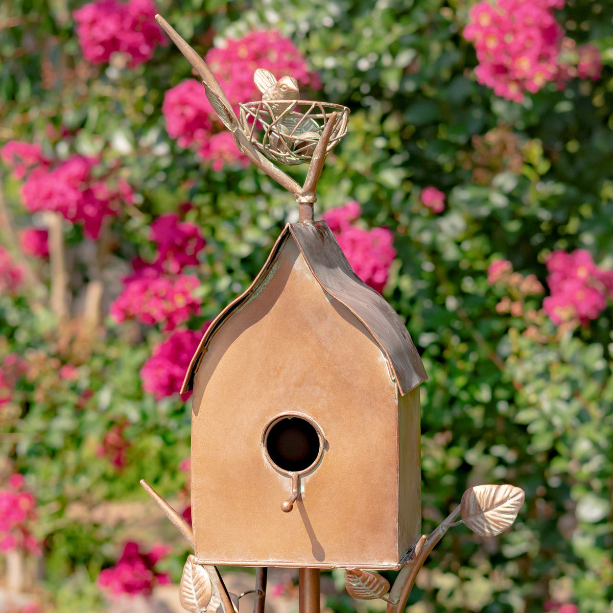 Front of a copper-toned birdhouse with a round entrance, sloped roof, and decorative bird and nest above.