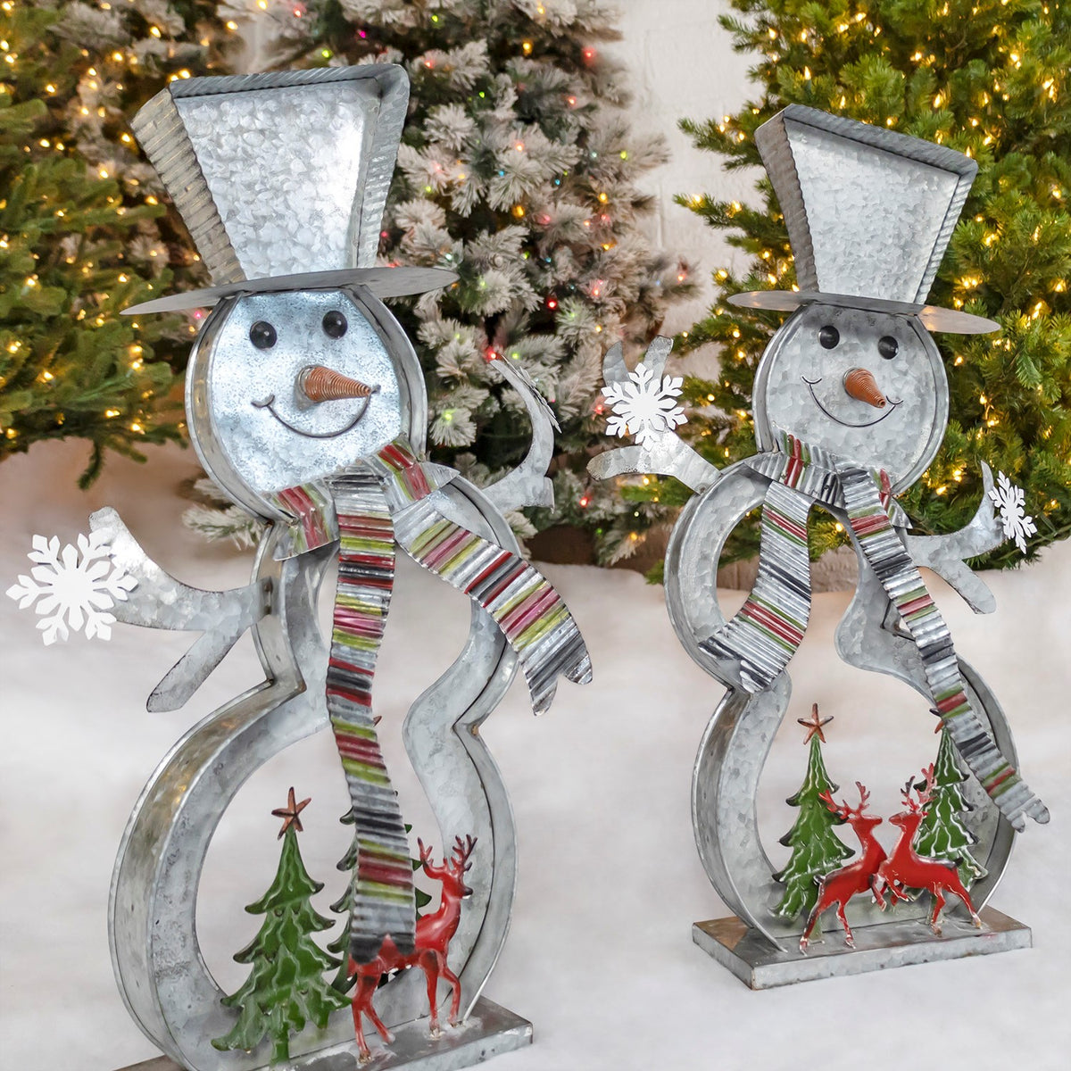 Two galvanized snowman decorations with striped scarves stand on bases beside painted trees and reindeer.