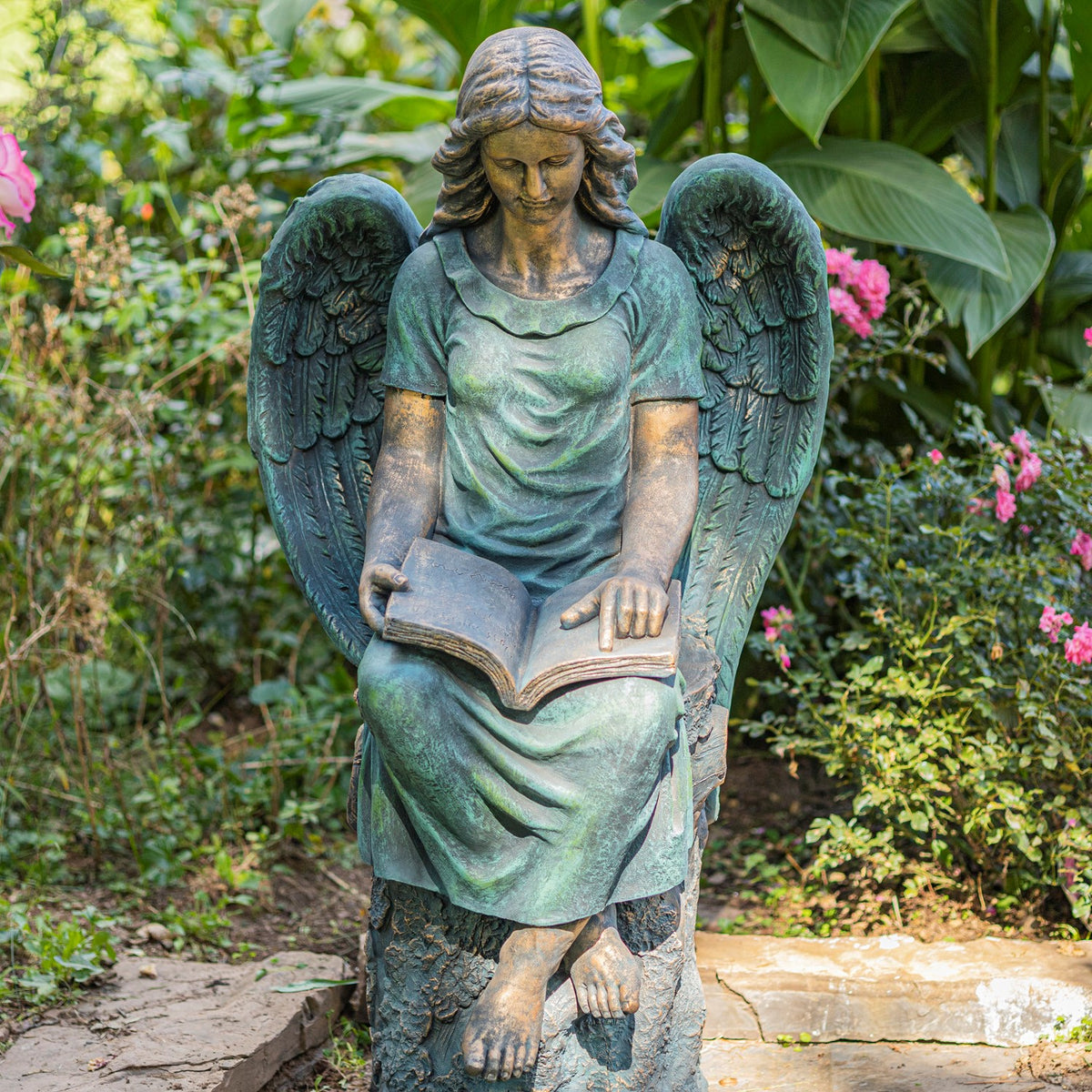 Full view of the seated angel statue reading an open book, with crossed feet and wings in a garden setting.