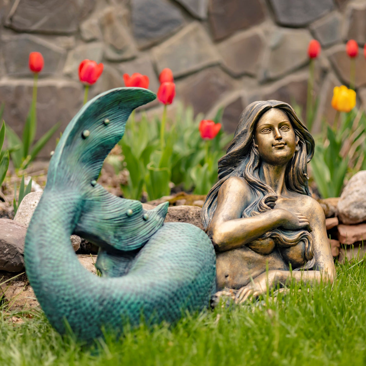 Lying mermaid garden statue with a curled teal tail beside a stone border and flowers.