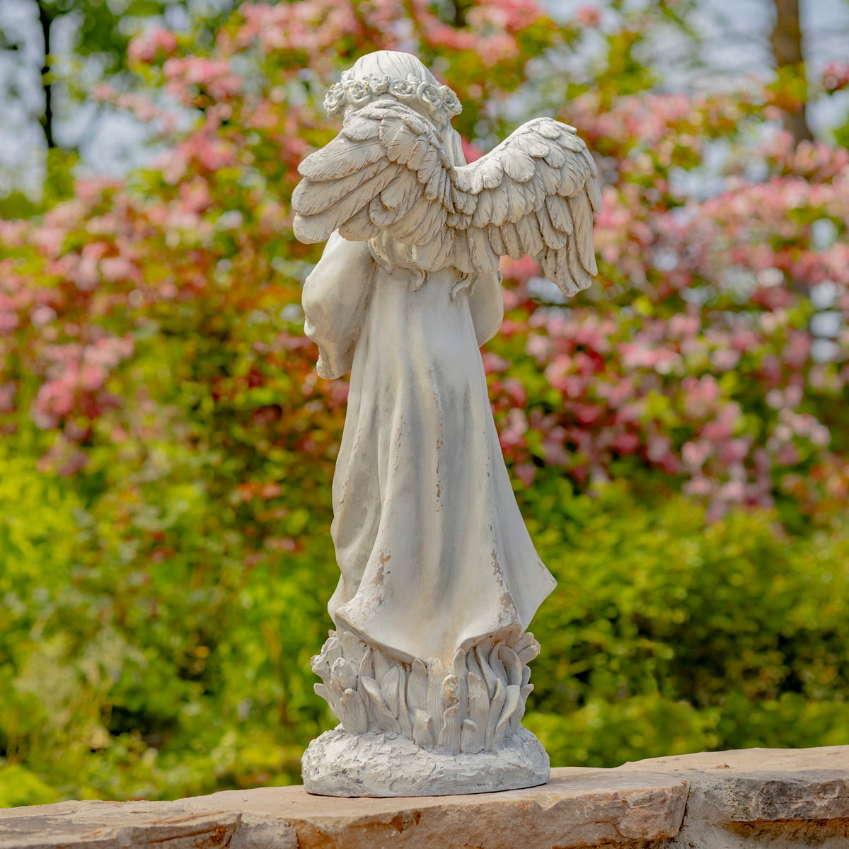 The angel statue viewed from behind, showing its layered wings, long robe, and flower-decorated base.