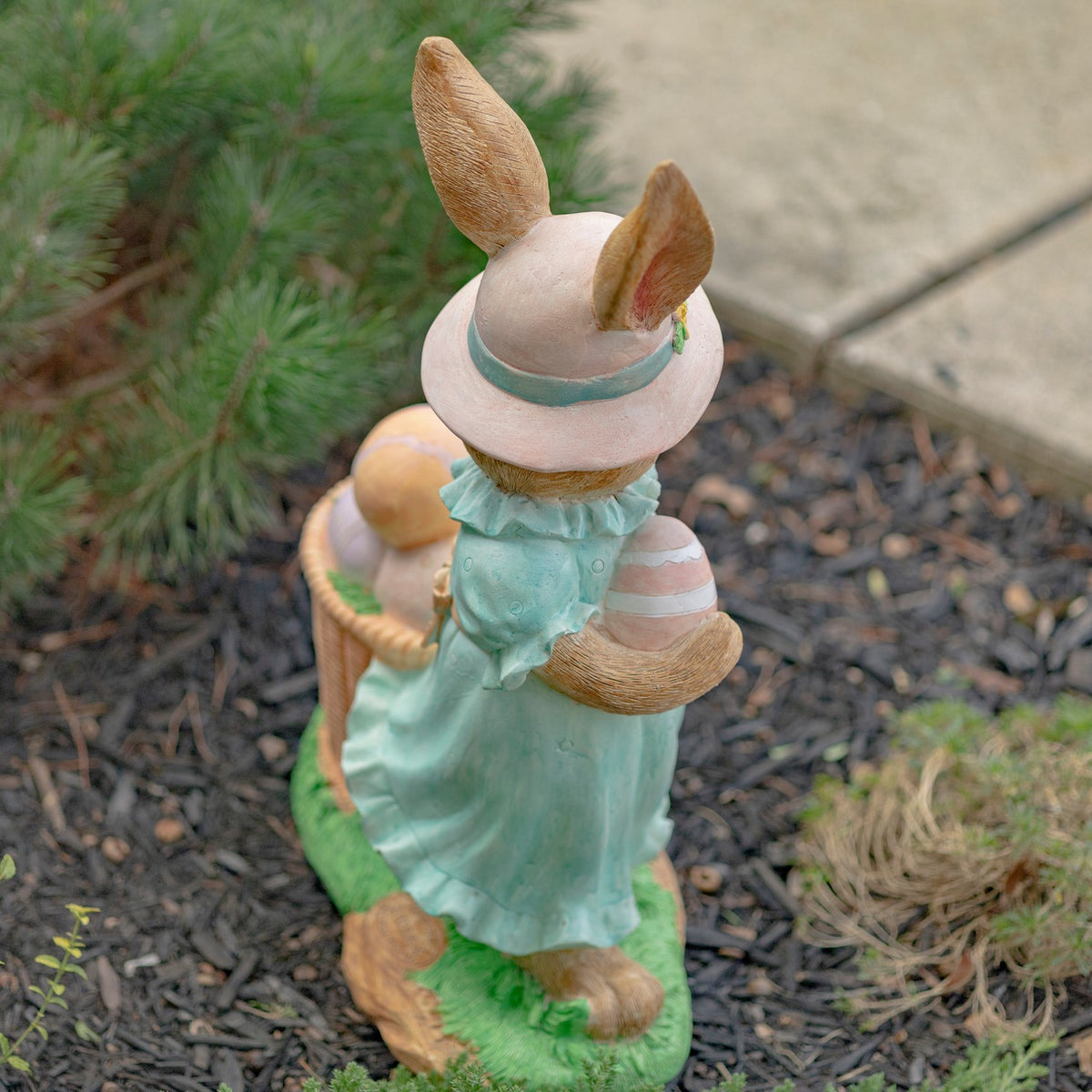 Back view of the rabbit statue in an aqua dress, standing on a grassy base beside an egg basket.