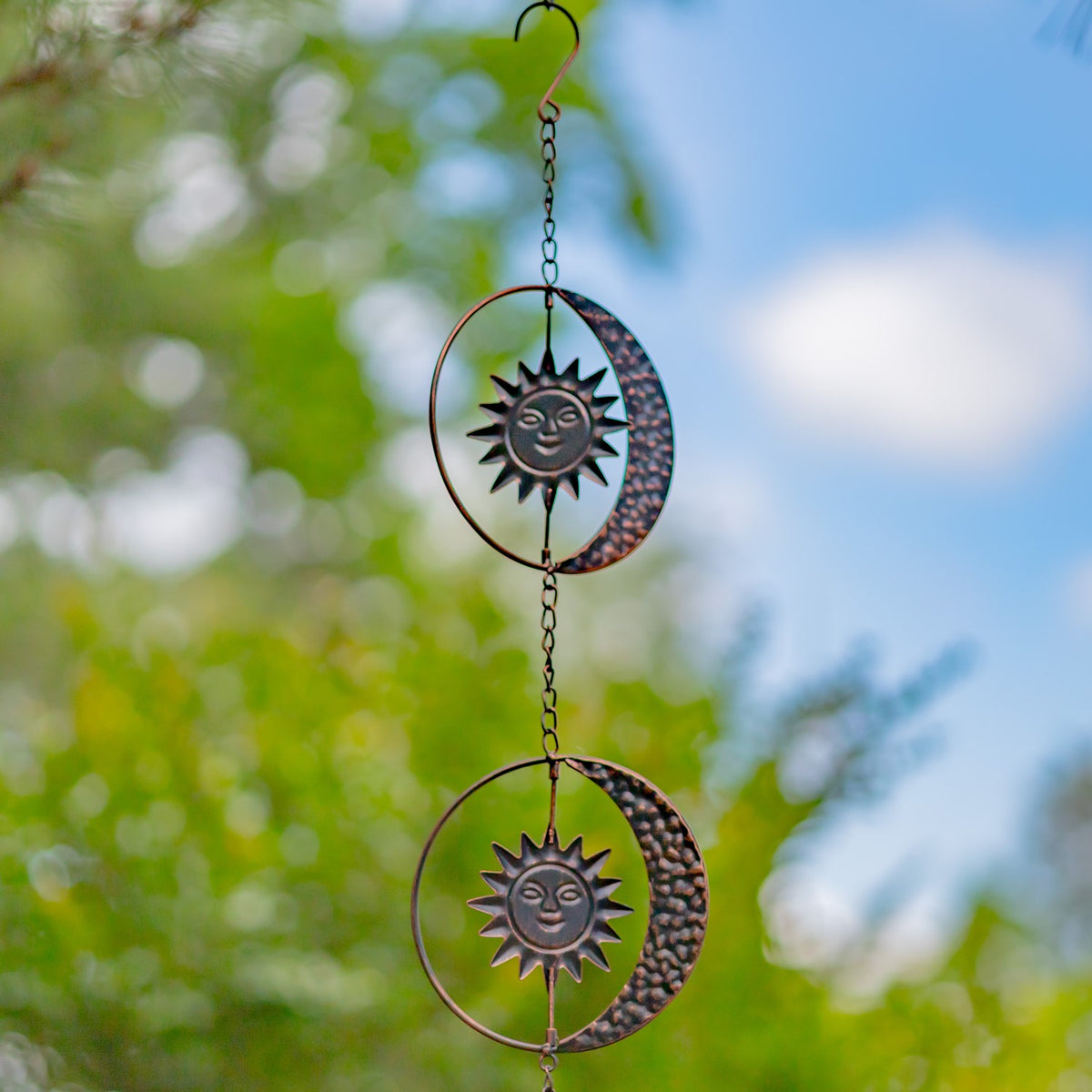 Antique bronze sun and crescent moon rain chain shown against sky and garden greenery