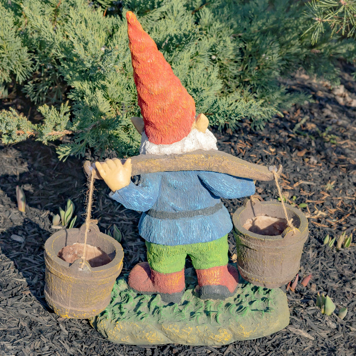 Back of the gnome carrying a wooden pole with two hanging buckets, standing among garden shrubs.