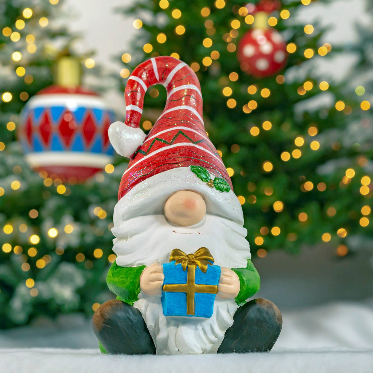 Seated gnome in a red candy cane hat holding a blue wrapped gift