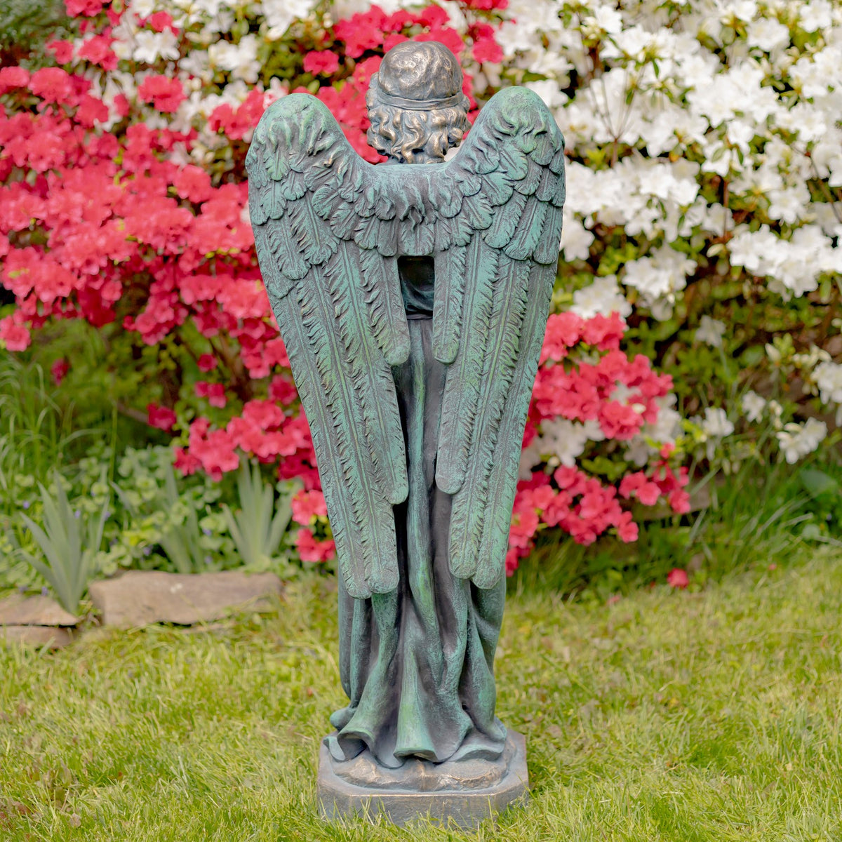 Full rear view of the angel statue, with long feathered wings and a flowing robe against a garden backdrop.