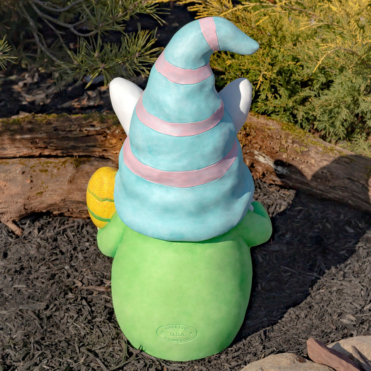 Rear view of the green gnome beneath a turquoise and pink striped hat outdoors