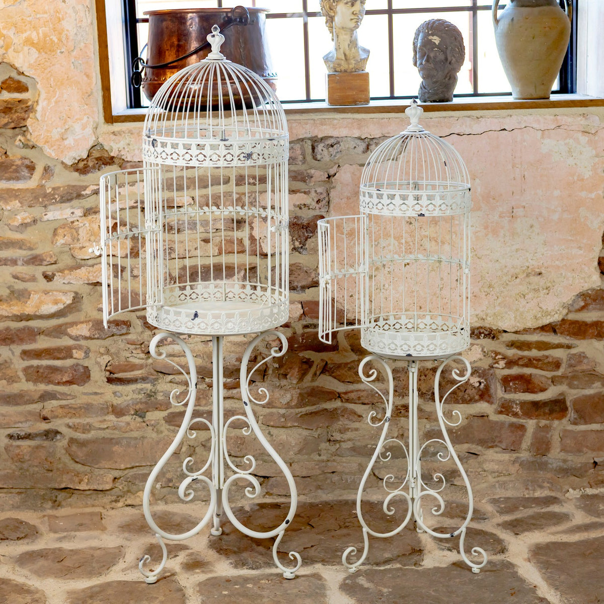 Two white iron birdcage planters with ornate scrollwork stands against a rustic stone wall.