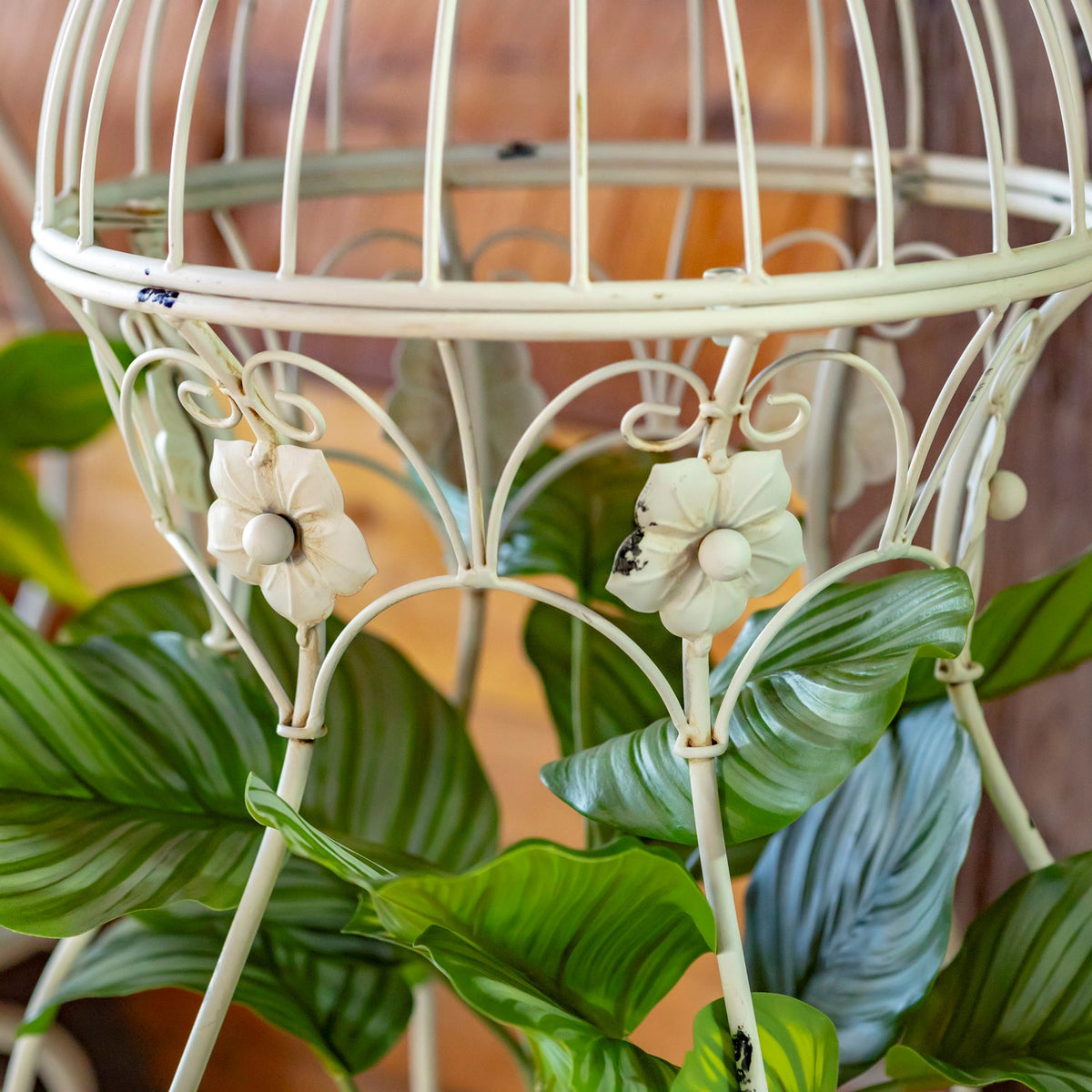 Close-up of pale metal birdcage bars and flower ornaments among green plant leaves.