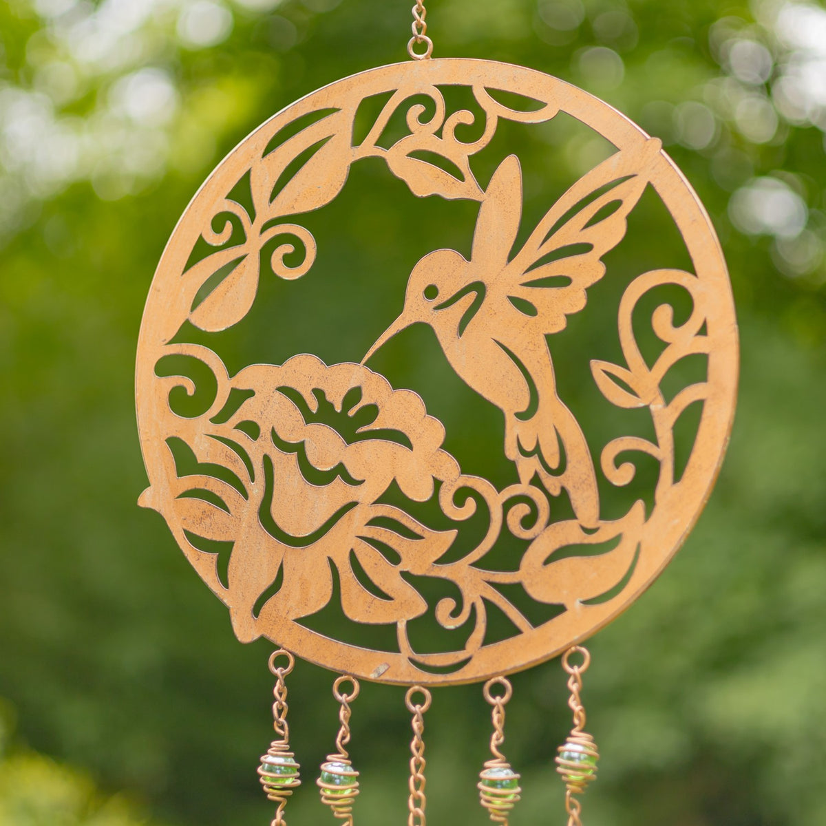 Copper-finish circular wind chime with a hummingbird and flowers against greenery