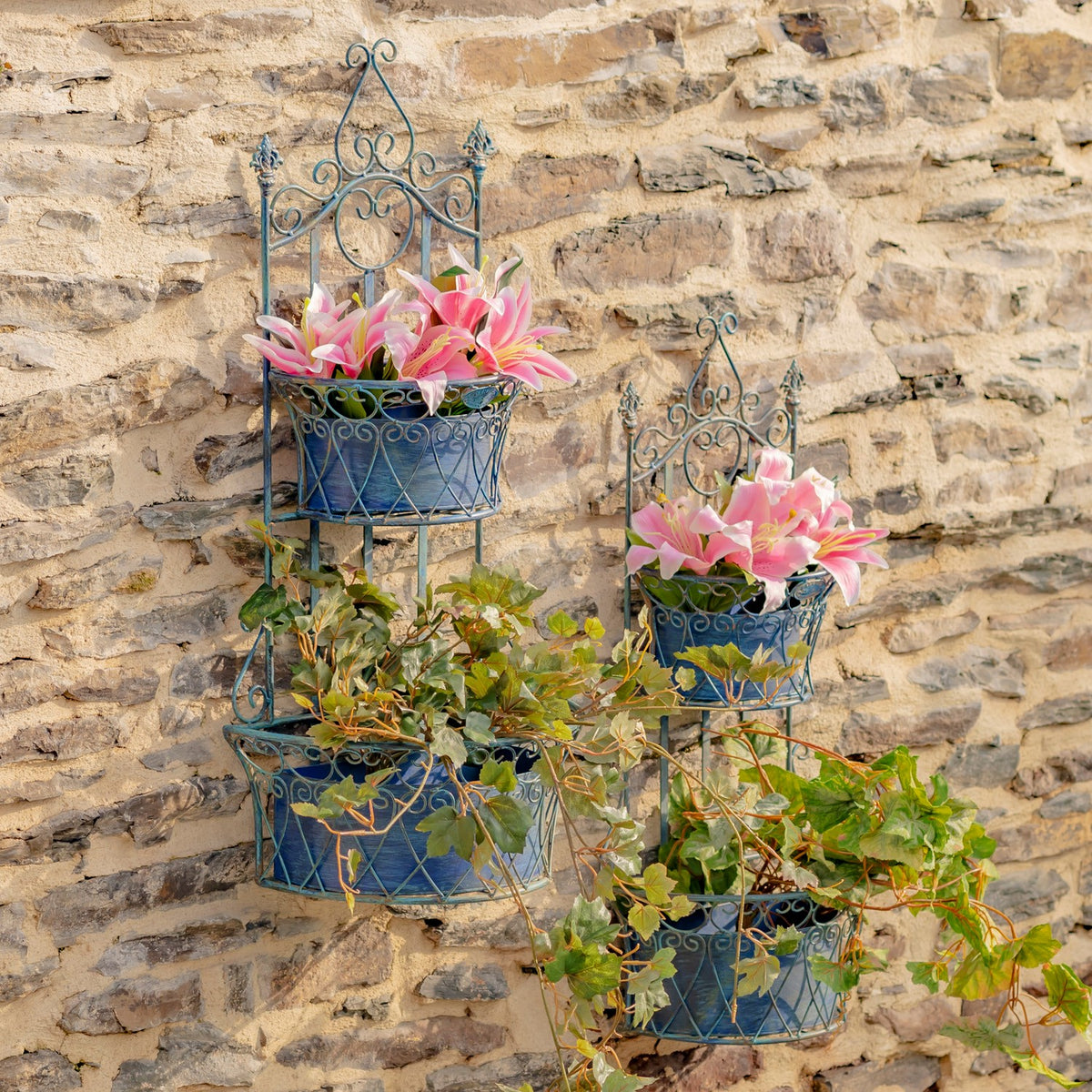 Two blue scrollwork wall planters with pink flowers and trailing greenery on a stone wall.