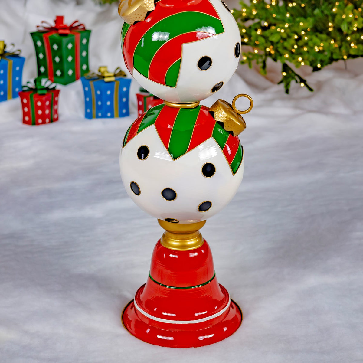 A red pedestal supports stacked red, green, and white ornament spheres with black dots beside wrapped gifts.