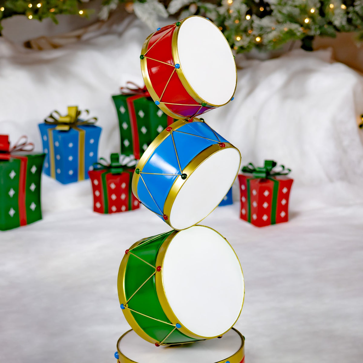 Three red, blue, and green drums stacked on a stand beside Christmas presents and a lit tree.