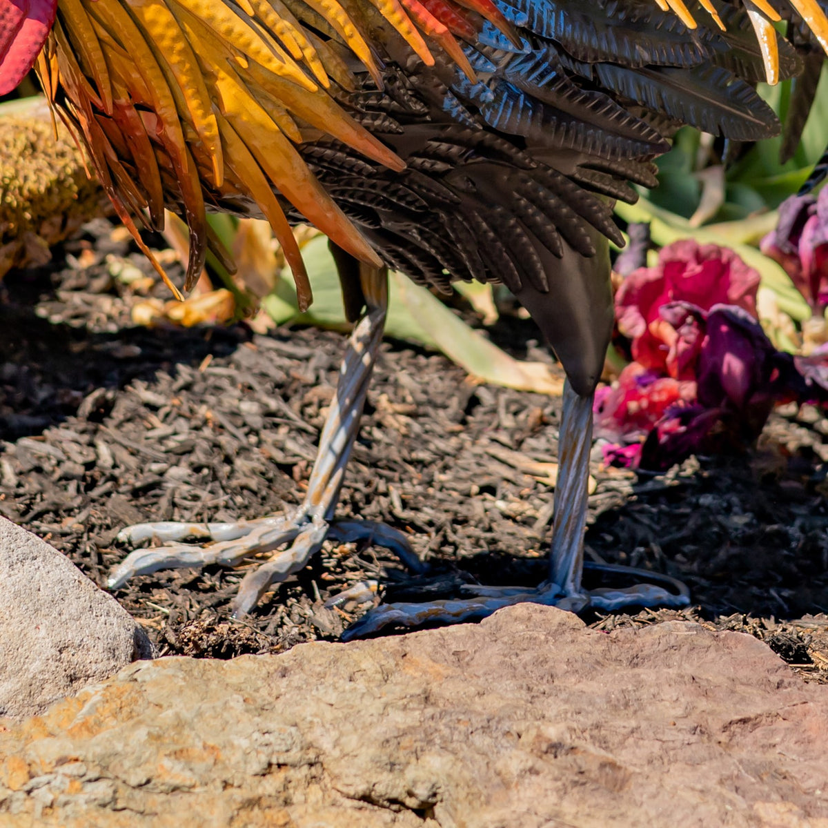 Close-up of the rooster’s slender metal legs and splayed feet on garden soil.