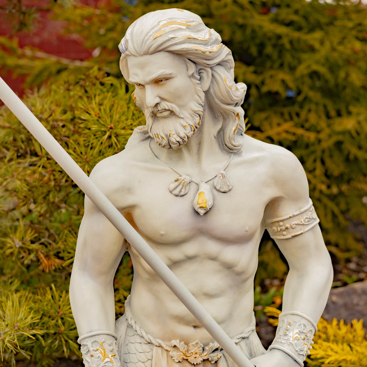 Upper-body view of a muscular merman statue holding a diagonal trident, with shell details and garden foliage behind.