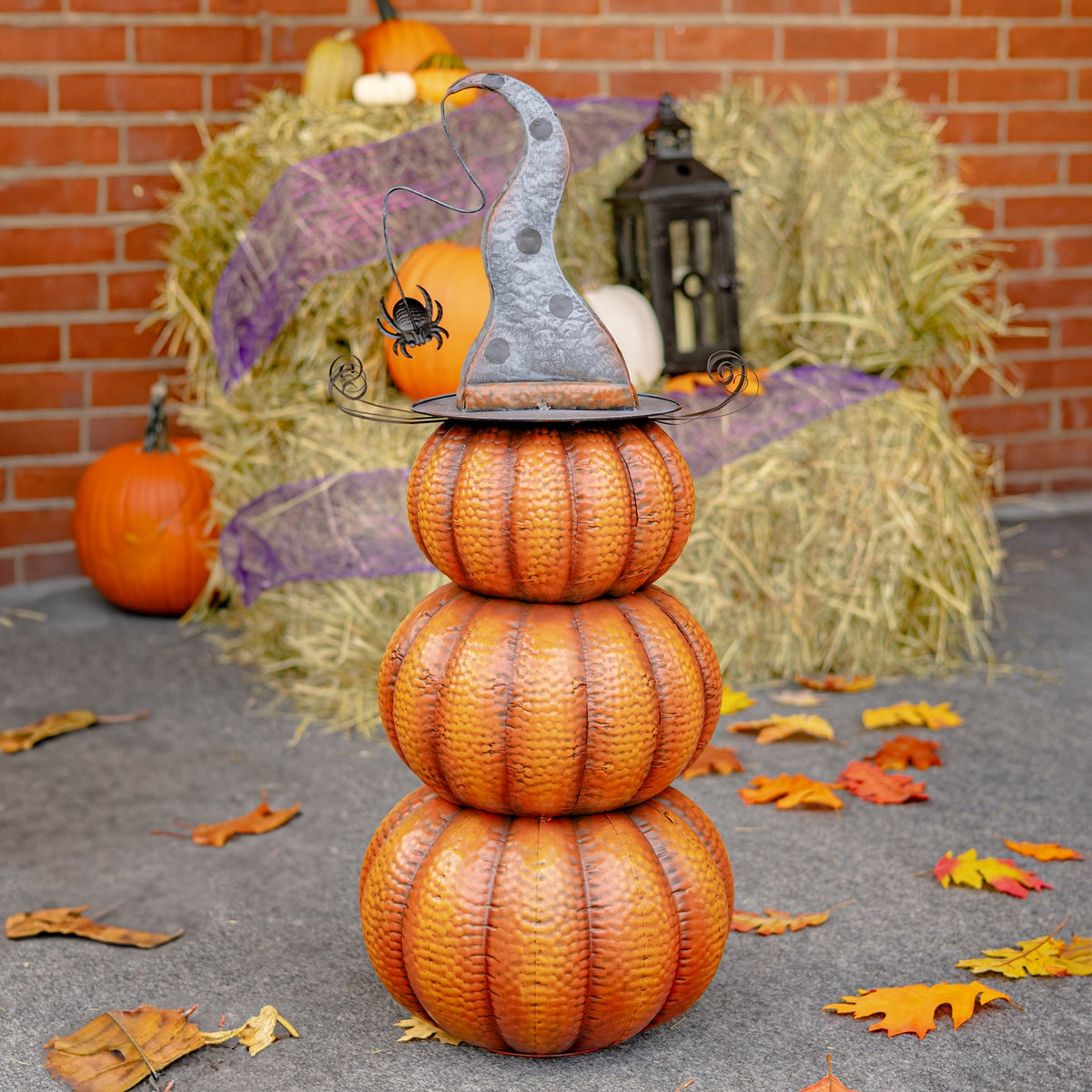 Three stacked orange pumpkins topped with a curled gray witch hat, staged among hay bales, leaves, and Halloween decor.