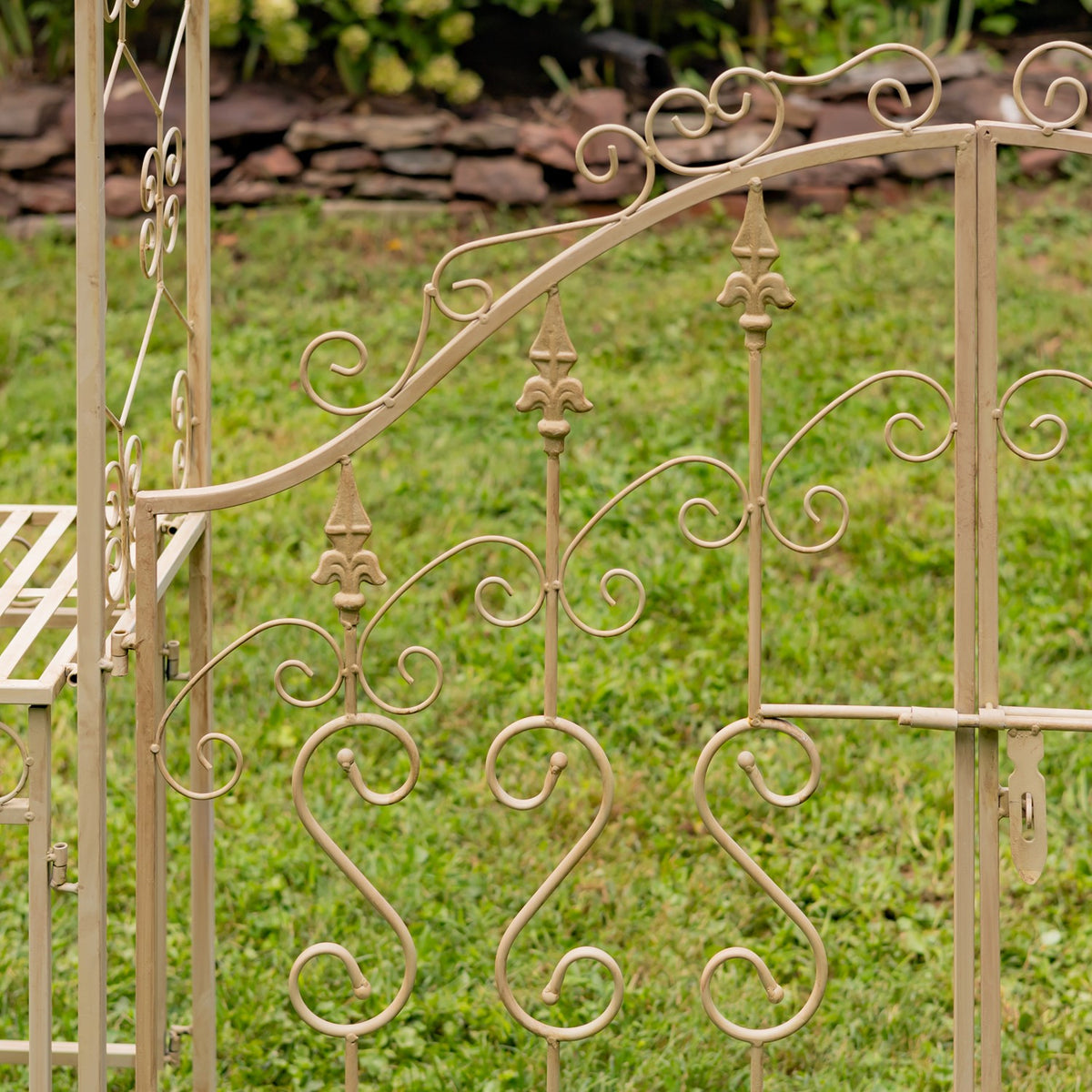Close view of curling metal scrollwork and fleur-de-lis accents on the garden archway, with part of a slatted stand at left.