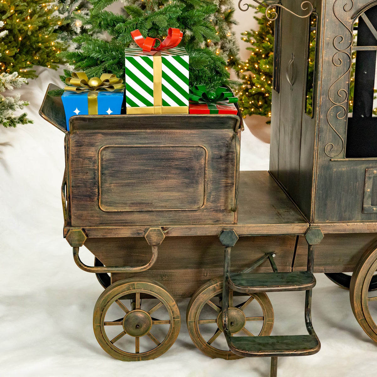 Close view of a dark metal train cart with gift boxes, spoked wheels and a small step beside a lit tree.