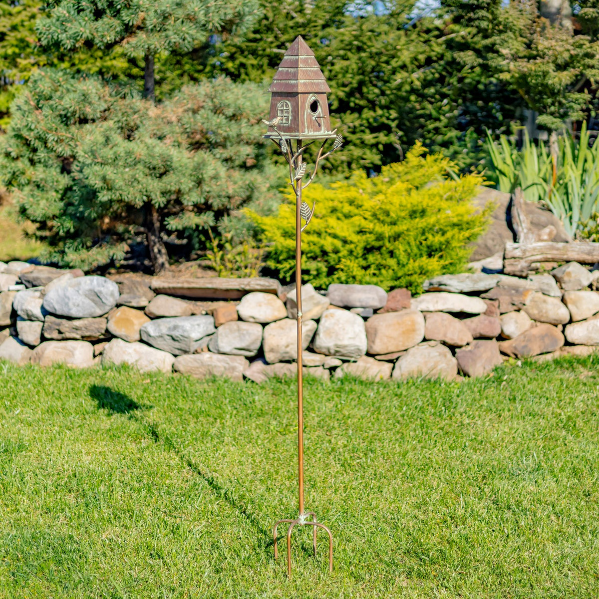 Full birdhouse stake viewed from a distance on a lawn beside a stone wall