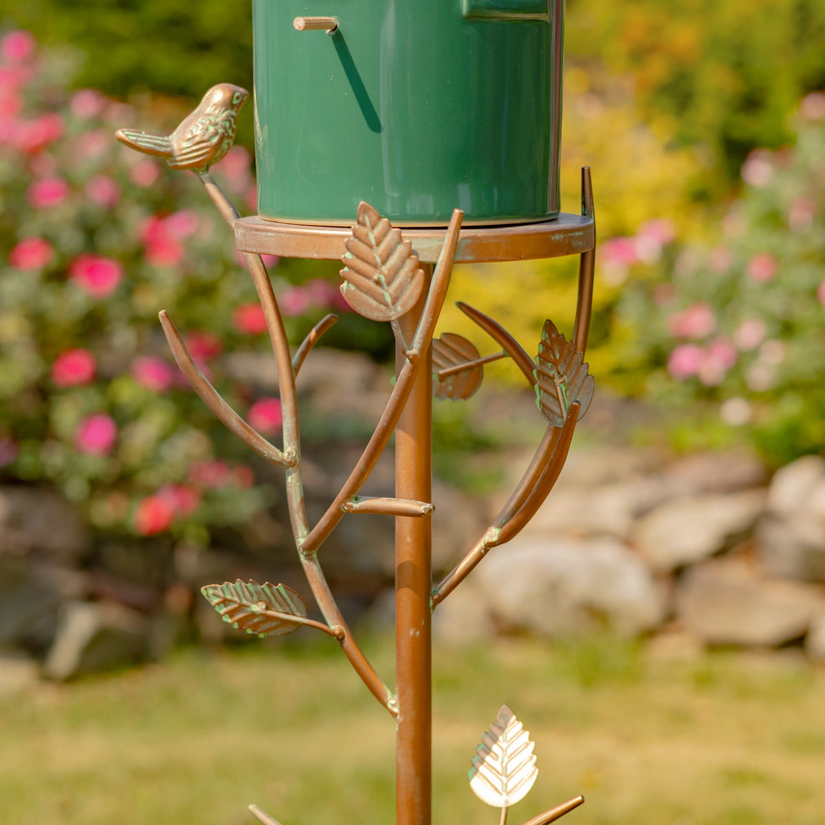 Green birdhouse mounted on a decorative brown stake with metal bird and leaf accents in a garden.